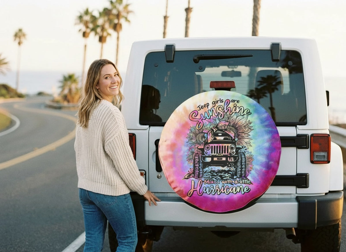 A woman beside a white Jeep whose spare tire cover carries the Jeep Girls Are Sunshine tie-dye design